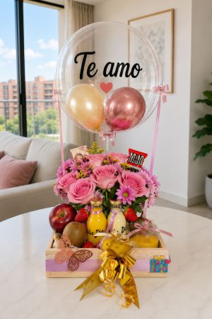 Clear balloon with the words 'Te amo' in black script, perched above a pink flower bouquet and a fruit and juice gift box on a table.